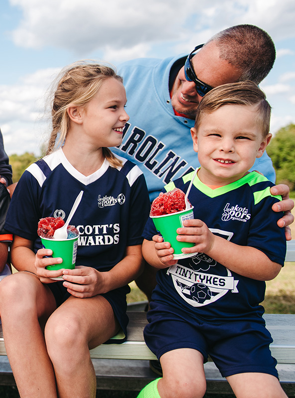 sports tournaments Two kids smile while holding shaved ice with their dad after a soccer tournament