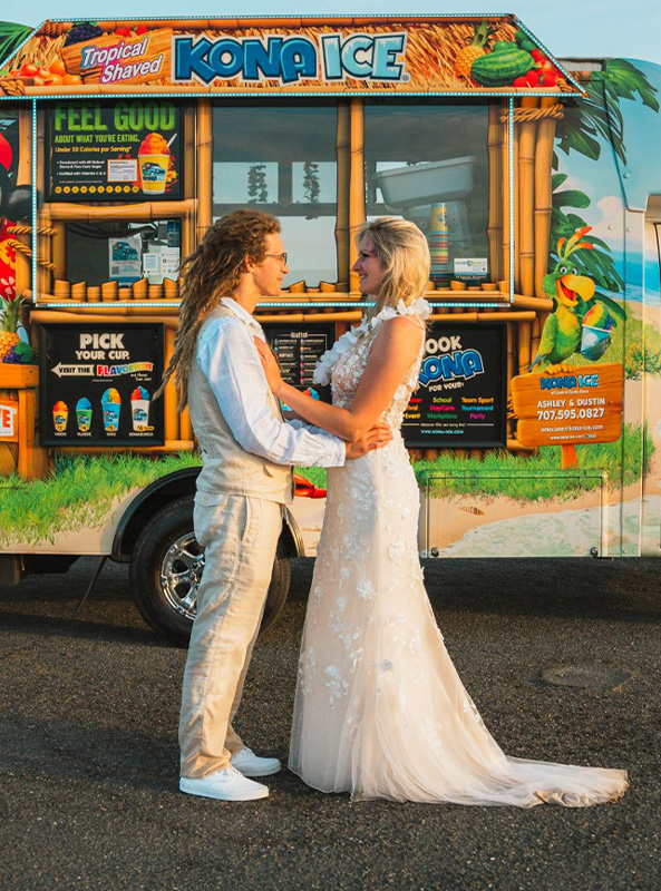 A little boy gives a thumbs up and holds a cup of red shaved ice as he stands in front of a Kona Ice truck that is catering a private event