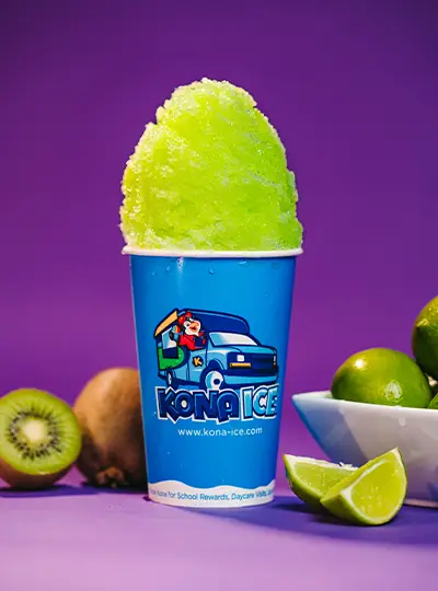 A blue Kona Ice cup filled with green shaved ice next to limes and kiwis 